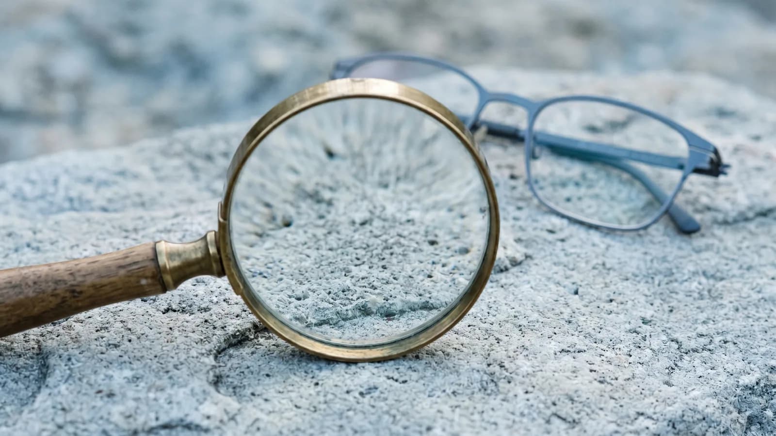 Ett förstoringsglas som ligger på en sval grå stenyta, med ett par tunna glasögon ur fokus i bakgrunden