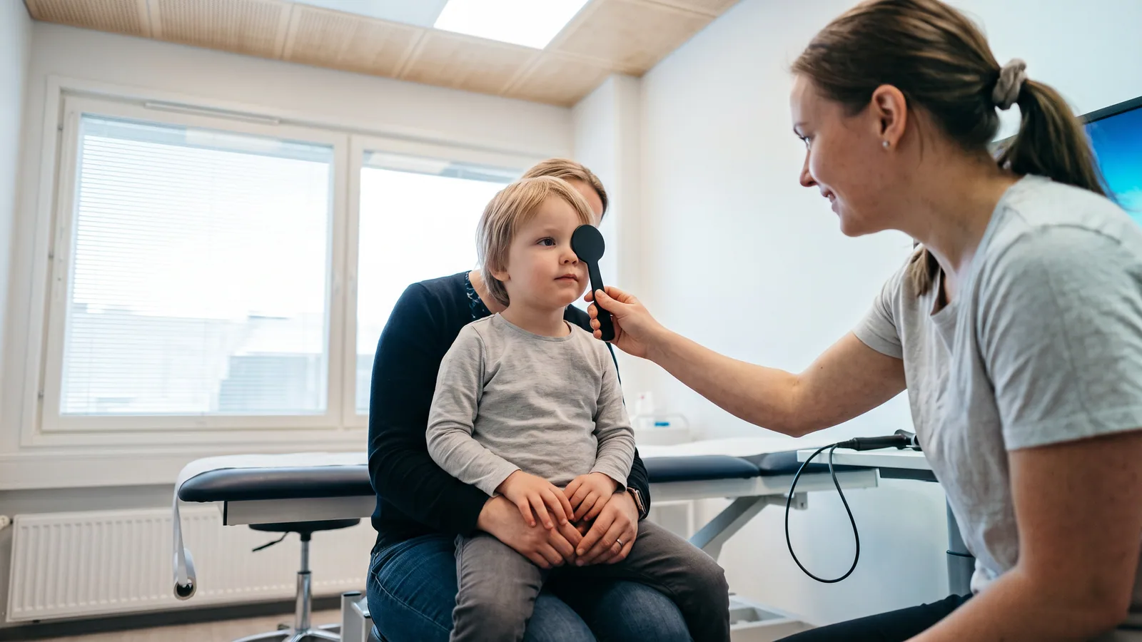 Ett barn med glasögon tittar uppmärksamt mot ett fönster i ett ljust nordiskt rum, det ena ögat lätt vänt inåt