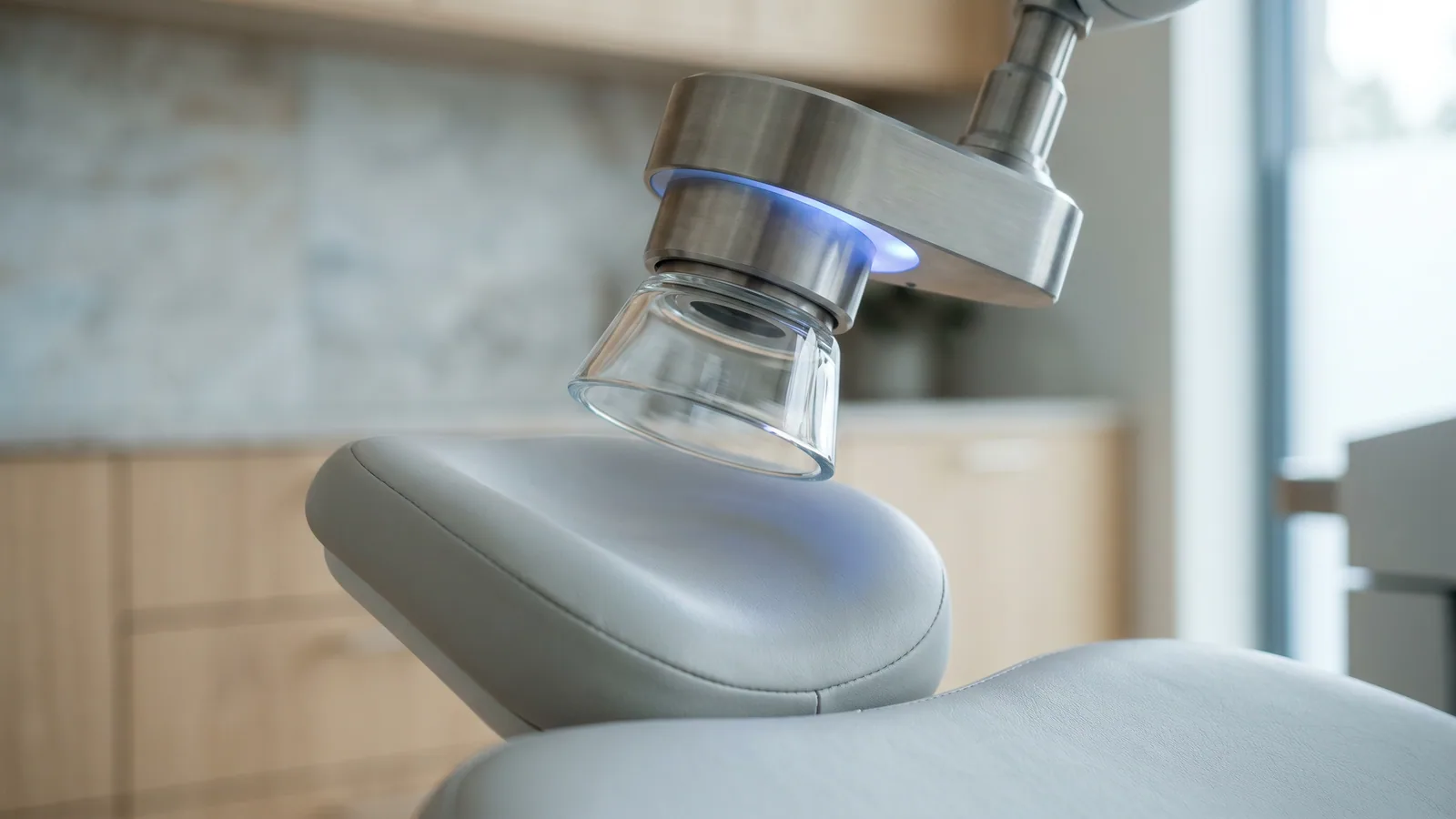 Närbild av dockningskonen på en femtosekundlaser för ögonlaser, med ett svagt blått ljus, ovanför en tom patientstol i en ljus nordisk ögonklinik.