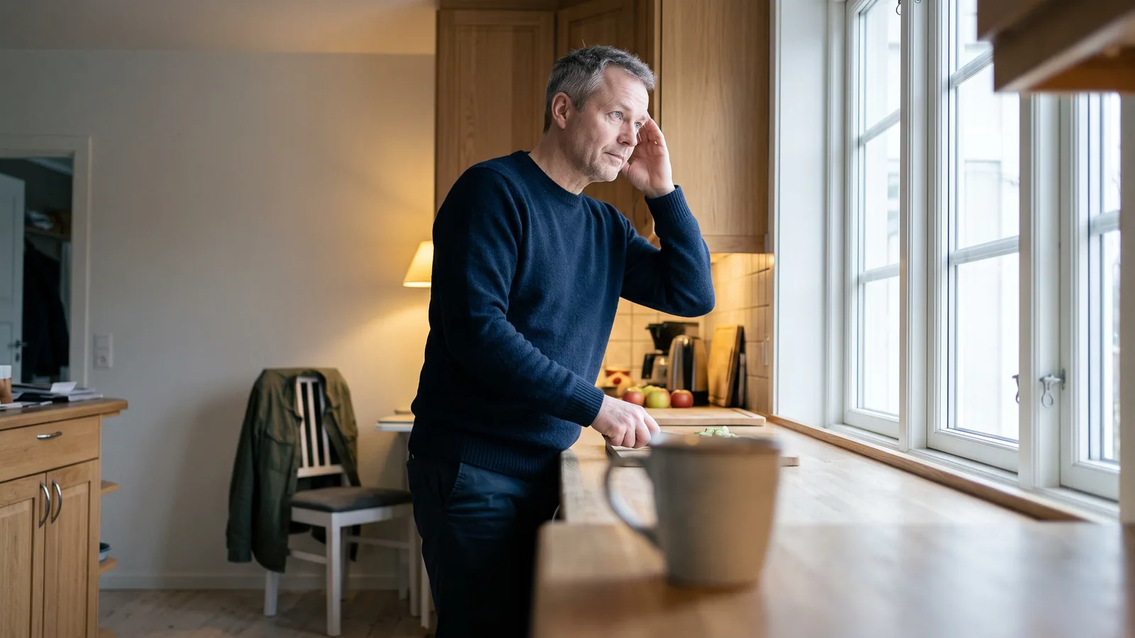 En man i femtioårsåldern står vid köksfönstret med handen mot tinningen och en frånvarande blick ut i det grå dagsljuset.
